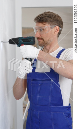 Professional male construction worker in a blue jumpsuit, safety goggles, and work gloves is screwing a screw into the wall using a cordless screwdriver, carrying out a careful home renovation Professional male construction worker in a blue jumpsuit, safety goggles, and work gloves is screwing a screw into the wall using a cordless screwdriver, carrying out a careful home renovation 132359291