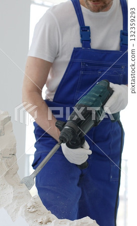Unknown male construction worker wearing blue overalls is demolishing white wall with rotary hammer drill, generating dust, close up horizontal view. Renovation Unknown male construction worker wearing blue overalls is demolishing white wall with rotary hammer drill, generating dust, close up horizontal view. Renovation 132359293