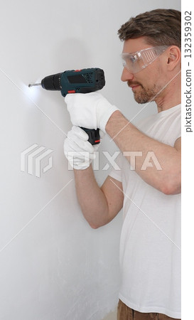 Middle aged man worker wearing white t-shirt and protective gloves, is drilling hole in a wall with cordless drill during home renovation work. Vertical portrait view Middle aged man worker wearing white t-shirt and protective gloves, is drilling hole in a wall with cordless drill during home renovation work. Vertical portrait view 132359302