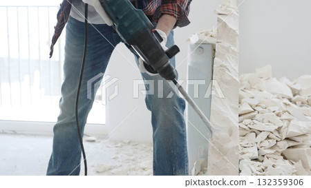 Close up of unknown male construction worker wearing red checkered shirt jeans and protective gloves, is demolishing white wall with rotary hammer drill, generating debris on the floor. Renovation Close up of unknown male construction worker wearing red checkered shirt jeans and protective gloves, is demolishing white wall with rotary hammer drill, generating debris on the floor. Renovation 132359306