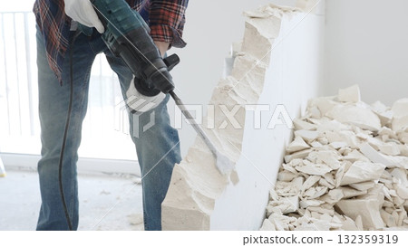 Close up of unknown male construction worker wearing red checkered shirt jeans and protective gloves, is demolishing white wall with rotary hammer drill, generating debris on the floor. Renovation 132359319