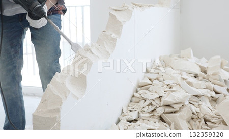 Close up of unknown male construction worker wearing red checkered shirt jeans and protective gloves, is demolishing white wall with rotary hammer drill, generating debris on the floor Close up of unknown male construction worker wearing red checkered shirt jeans and protective gloves, is demolishing white wall with rotary hammer drill, generating debris on the floor 132359320