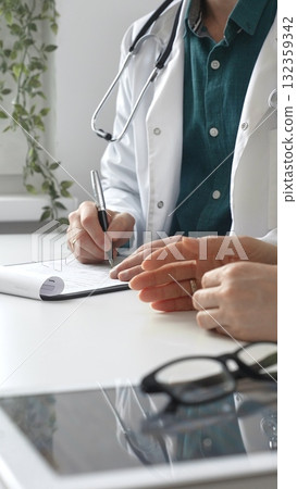 Doctor man wearing medical coat over a green shirt is listening to patient and writing a prescription during a consultation in a modern clinic. Medicine and health care concept Doctor man wearing medical coat over a green shirt is listening to patient and writing a prescription during a consultation in a modern clinic. Medicine and health care concept 132359342