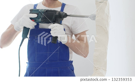 Close up of unrecognizable male construction worker wearing blue overalls and protective white gloves, is demolishing white wall with rotary hammer drill, generating dust, horizontal view. Renovation 132359353
