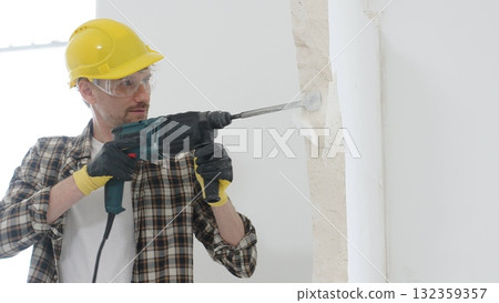 Portrait of a man construction worker wearing beige checkered shirt , yellow hard hat and protective gloves, is demolishing white wall with rotary hammer drill, generating dust. Renovation concept 132359357