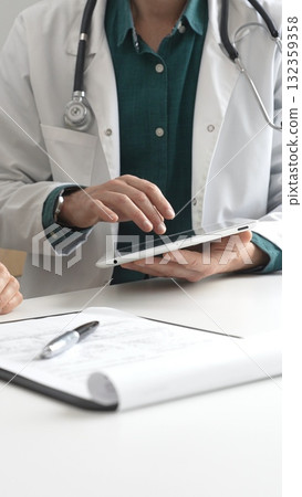 Male doctor in lab coat over a green shirt, and stethoscope gesturing while explaining diagnosis to patient using tablet in modern medical office. Medicine and health care concept 132359358