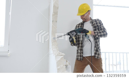 Man construction worker wearing beige checkered shirt , yellow hard hat and protective gloves, is demolishing white wall with rotary hammer drill, generating dust, medium long shot. Renovation concept 132359359