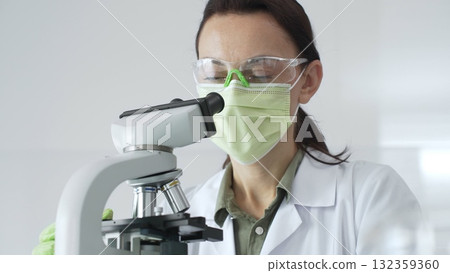 Female professional scientist examining specimen, placing slide under microscope while wearing green protective gear in laboratory workspace. Medicine, health care and science concept Female professional scientist examining specimen, placing slide under microscope while wearing green protective gear in laboratory workspace. Medicine, health care and science concept 132359360
