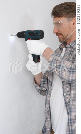 Middle aged male professional worker wearing grey checked shirt and white gloves, is drilling hole in a wall with cordless drill during home renovation work. Vertical portrait view 132359382