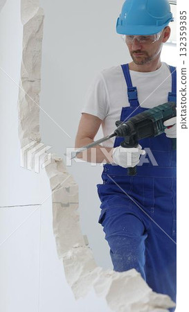 Male construction worker wearing blue overalls, protective helmet and white gloves, is demolishing white wall with rotary hammer drill, generating dust, close up horizontal view Male construction worker wearing blue overalls, protective helmet and white gloves, is demolishing white wall with rotary hammer drill, generating dust, close up horizontal view 132359385