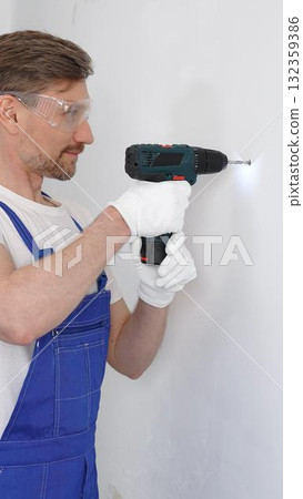Professional male construction worker in a blue jumpsuit, safety goggles, and work gloves is screwing a screw into the wall using a cordless screwdriver. Home renovation concept Professional male construction worker in a blue jumpsuit, safety goggles, and work gloves is screwing a screw into the wall using a cordless screwdriver. Home renovation concept 132359386