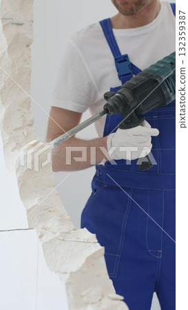 Unknown male construction worker wearing blue overalls and protective white gloves, is demolishing white wall with rotary hammer drill, generating dust, close up horizontal view. Renovation Unknown male construction worker wearing blue overalls and protective white gloves, is demolishing white wall with rotary hammer drill, generating dust, close up horizontal view. Renovation 132359387