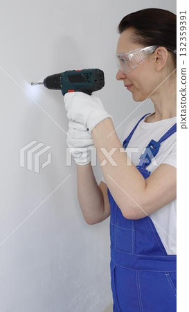Professional female worker wearing blue overall, safety glasses and gloves, is using cordless drill to make hole in white wall during renovation work. Portrait view 132359391