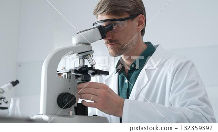 Adult male scientist wearing lab coat and protective glasses using microscope, doing scientific research in modern laboratory. Medicine, healthcare and science concept Adult male scientist wearing lab coat and protective glasses using microscope, doing scientific research in modern laboratory. Medicine, healthcare and science concept 132359392