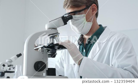 Adult male scientist wearing white mask, lab coat and protective glasses using microscope, doing scientific research in modern laboratory. Medicine, health care and science concept Adult male scientist wearing white mask, lab coat and protective glasses using microscope, doing scientific research in modern laboratory. Medicine, health care and science concept 132359404