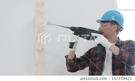 Male construction worker wearing red checkered shirt, blue protective helmet and gloves, is demolishing white wall with rotary hammer drill, generating dust, portrait view. Renovation concept 132359421
