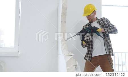 Male construction worker wearing beige checkered shirt, yellow hard hat and protective gloves, is demolishing white wall with rotary hammer drill, generating dust, medium long shot. Renovation project 132359422