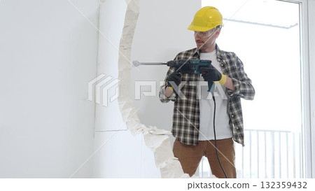 Male construction worker wearing beige checkered shirt, yellow hard hat and protective gloves, is demolishing white wall with rotary hammer drill, generating dust, medium long shot. Renovation 132359432