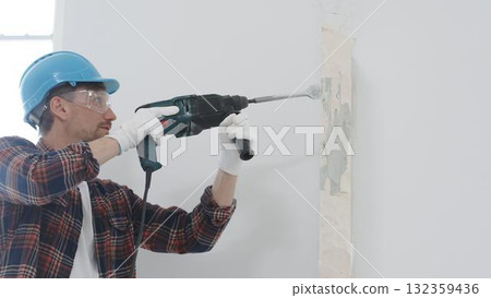 Man construction worker wearing red checkered shirt, blue hard hat and protective gloves, is demolishing white wall with rotary hammer drill, generating dust, portrait view. Renovation concept Man construction worker wearing red checkered shirt, blue hard hat and protective gloves, is demolishing white wall with rotary hammer drill, generating dust, portrait view. Renovation concept 132359436
