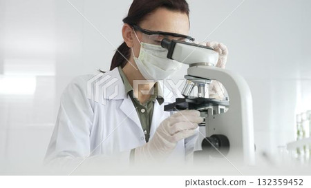 Female scientist wearing a lab coat, face mask, and safety glasses carefully adjusts a microscope, conducting research in a brightly lit laboratory setting. Medicine, healthcare and science concept Female scientist wearing a lab coat, face mask, and safety glasses carefully adjusts a microscope, conducting research in a brightly lit laboratory setting. Medicine, healthcare and science concept 132359452