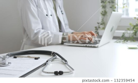 Stethoscope and medical records are on the desk opposite the doctor is typing on a laptop with a in a medical office, suggesting online healthcare or telemedicine services. Medicine concept Stethoscope and medical records are on the desk opposite the doctor is typing on a laptop with a in a medical office, suggesting online healthcare or telemedicine services. Medicine concept 132359454