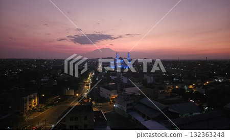 A magnificent mosque, dramatically illuminated in striking blue light against a vibrant purple and orange twilight sky, stands prominently in a sprawling city landscape with distant majestic mountains 132363248