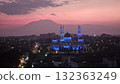 A magnificent mosque, dramatically illuminated in striking blue light against a vibrant purple and orange twilight sky, stands prominently in a sprawling city landscape with distant majestic mountains 132363249