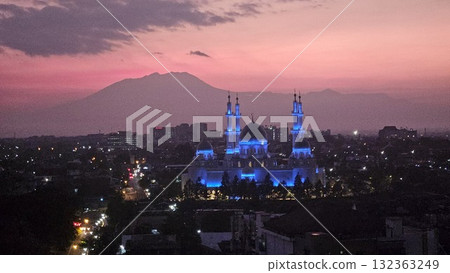 A magnificent mosque, dramatically illuminated in striking blue light against a vibrant purple and orange twilight sky, stands prominently in a sprawling city landscape with distant majestic mountains A magnificent mosque, dramatically illuminated in striking blue light against a vibrant purple and orange twilight sky, stands prominently in a sprawling city landscape with distant majestic mountains 132363249