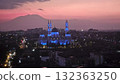 A magnificent mosque, dramatically illuminated in striking blue light against a vibrant purple and orange twilight sky, stands prominently in a sprawling city landscape with distant majestic mountains 132363250