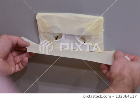 Close-up of the hands, a man glues the outlet with sticky tape to protect it from paint during repairs. 132364117