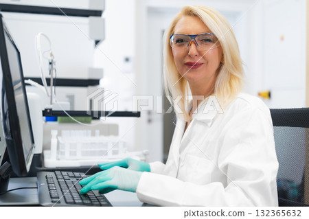 Blonde woman scientist in laboratory working on computer in front of high performance liquid chromatography HPLC equipment Blonde woman scientist in laboratory working on computer in front of high performance liquid chromatography HPLC equipment 132365632