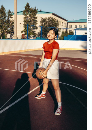 Teenage girl stands on a basketball court holding a ball. She has dark hair, wears a red shirt and white shorts, and smiles in the sunlight. 132366900