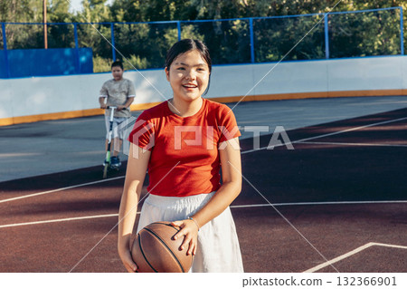 A young Asian girl smiles while holding a basketball on an outdoor court. A boy rides a scooter in the background. The scene captures fitness and youth activities. 132366901
