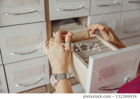 A person with light skin and short brown hair organizes index cards in a wooden drawer. The setting is a modern library or coworking space. 132366934
