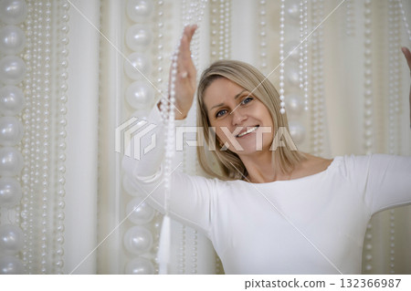 Smiling middle-aged Caucasian woman with long blonde hair, wearing a white top, surrounded by decorative pearl strands, celebrating Christmas with a magical atmosphere. Smiling middle-aged Caucasian woman with long blonde hair, wearing a white top, surrounded by decorative pearl strands, celebrating Christmas with a magical atmosphere. 132366987