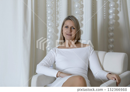 A young Caucasian woman with blond hair sits on a white chair. She wears a white dress and smiles, surrounded by a festive, magical Christmas backdrop. A young Caucasian woman with blond hair sits on a white chair. She wears a white dress and smiles, surrounded by a festive, magical Christmas backdrop. 132366988