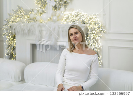 A young Caucasian woman with blonde hair sits on a white sofa. She wears a white off-shoulder dress. The background features festive Christmas decorations and soft lights. 132366997