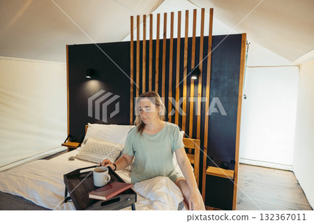 A young Caucasian woman with brown hair sits on a bed in a glamping tent. She holds a cup and has a book on a tray beside her. The interior features wooden accents. A young Caucasian woman with brown hair sits on a bed in a glamping tent. She holds a cup and has a book on a tray beside her. The interior features wooden accents. 132367011