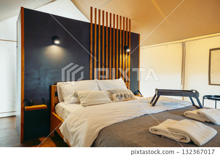 A cozy glamping tent interior featuring a comfortable bed with white linens, wooden accents, and soft lighting. The space is designed for relaxation and comfort. 132367017
