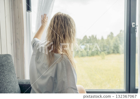 A young Caucasian woman with wavy blonde hair sits on a sofa, pulling back a curtain to reveal a green landscape outside a window. A young Caucasian woman with wavy blonde hair sits on a sofa, pulling back a curtain to reveal a green landscape outside a window. 132367067