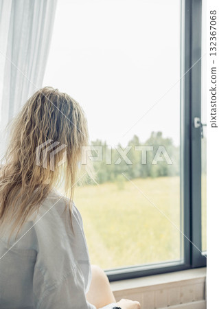 A young Caucasian woman with wavy hair sits by a large window, gazing at a green field outside. The scene conveys a sense of relaxation and escape. A young Caucasian woman with wavy hair sits by a large window, gazing at a green field outside. The scene conveys a sense of relaxation and escape. 132367068