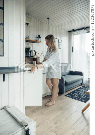 A young Caucasian woman with long brown hair stands in a cozy vacation home, reaching for items in a white cabinet. Natural light fills the room, creating a warm atmosphere. A young Caucasian woman with long brown hair stands in a cozy vacation home, reaching for items in a white cabinet. Natural light fills the room, creating a warm atmosphere. 132367072
