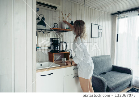 A young Caucasian woman with long brown hair stands in a cozy cabin kitchen, reaching for a cup. The space is bright and inviting, perfect for a vacation escape. A young Caucasian woman with long brown hair stands in a cozy cabin kitchen, reaching for a cup. The space is bright and inviting, perfect for a vacation escape. 132367073
