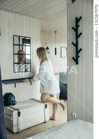 A young Caucasian woman with long blond hair stands in a cozy vacation cabin. She wears a white shirt and looks into a mirror, reflecting a serene escape atmosphere. A young Caucasian woman with long blond hair stands in a cozy vacation cabin. She wears a white shirt and looks into a mirror, reflecting a serene escape atmosphere. 132367076