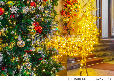 Decorated Christmas or New Year's tree, adorned with Christmas tree ornaments and garlands that glow and sparkle, creating bokeh effect, next to a figure of a deer and two figures of gingerbread men 132372975