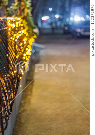 An iron black fence decorated with Christmas garlands stretching the entire length of the street, blurred yellow bulbs create a pleasant, beautiful bokeh effect. Seasonal Holiday Decorations An iron black fence decorated with Christmas garlands stretching the entire length of the street, blurred yellow bulbs create a pleasant, beautiful bokeh effect. Seasonal Holiday Decorations 132372976
