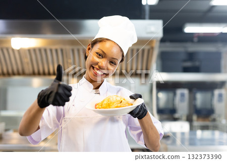 Enthusiastic women chef poses with plate dish of food in kitchen. Female wears chef hat and gloves, smiling and giving thumbs up, showing satisfaction in meal has prepared. Enthusiastic women chef poses with plate dish of food in kitchen. Female wears chef hat and gloves, smiling and giving thumbs up, showing satisfaction in meal has prepared. 132373390