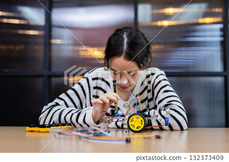 Student girl concentrates on assembling robotic car with wires and components at workshop. Engineering classroom with technology and enhancing creativity and learning. STEM education concept. 132373409