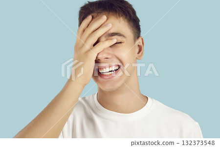 Portrait of young smiling boy covering his eyes with hand isolated on a blue background. 132373548