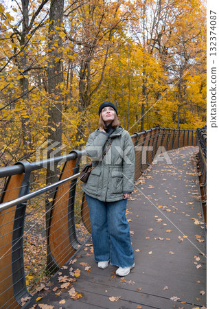 Woman in a green quilted jacket and black knit hat walking on the wooden eco trail bridge at VDNH park, surrounded by golden autumn trees 132374087